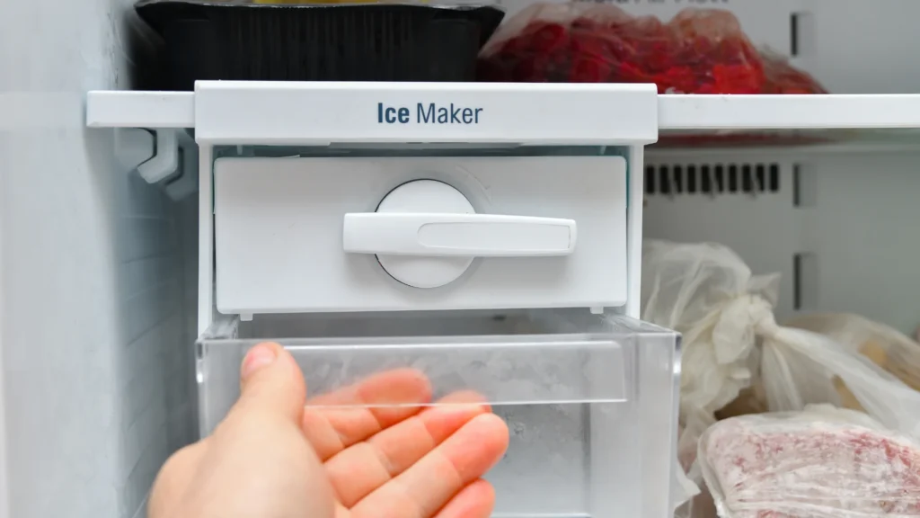 Hand beneath a manual twist-style ice tray and collection bin in a freezer